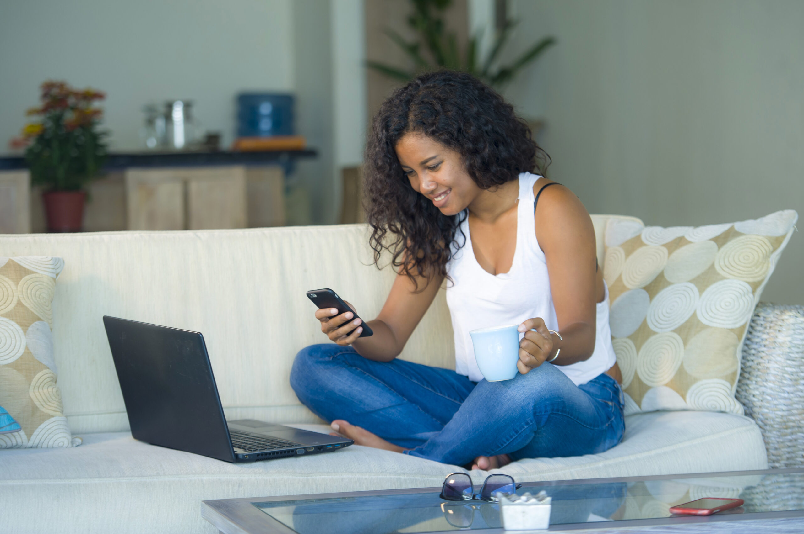 lifestyle portrait of young happy and beautiful black afro American woman using internet texting on mobile phone while working on laptop computer at home living room sofa couch relaxed